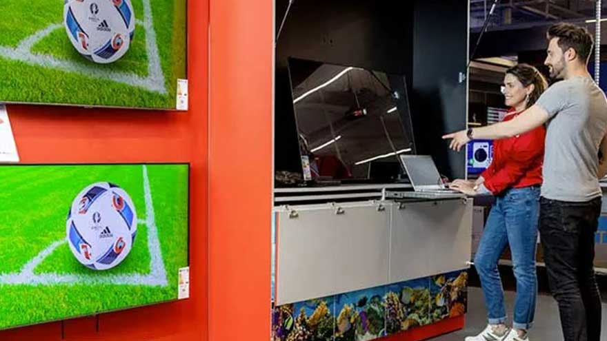 Twee tv's tonen een voetbal op groen gras tegen een oranje muur. Een laptop staat op een toonbank.