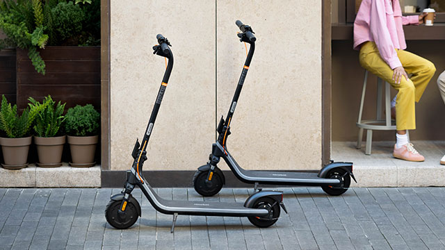 Twee zwarte elektrische steps staan op een grijze stenen bestrating. Achter de steps zijn planten in potten en een beige muur te zien.