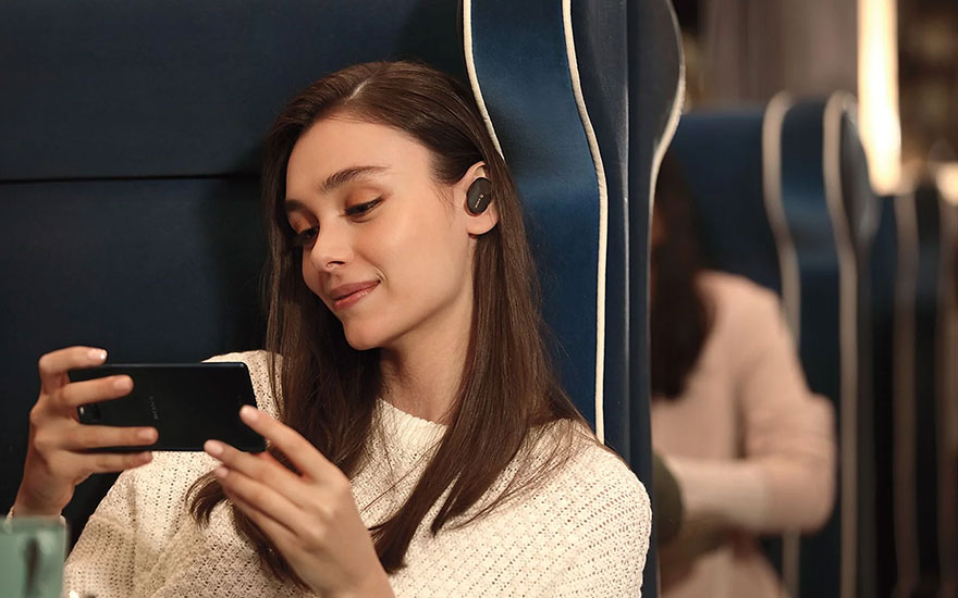 Dans un train, une personne utilise un smartphone noir, des oreillettes noires dans les oreilles, assise sur un siège bleu.