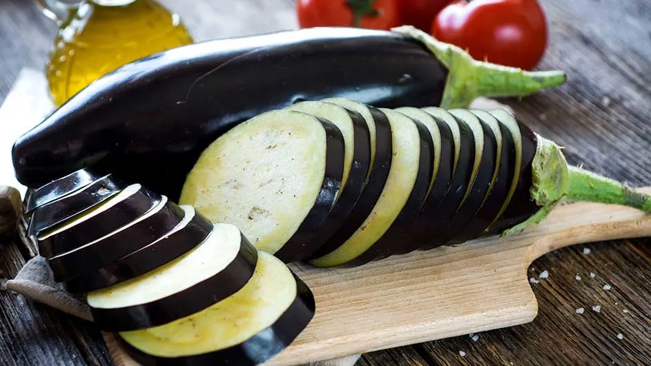 Een close-up van een aubergine, in plakjes gesneden, op een houten plank met olie en tomaten op de achtergrond.