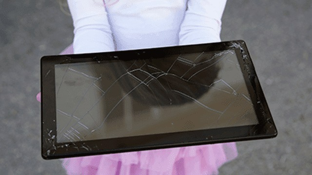 Une tablette noire est tenue. L'écran est fissuré. Le fond est gris.