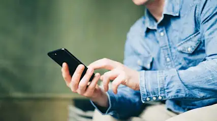 Een persoon met een blauw denim hemd gebruikt een zwarte smartphone tegen een groene achtergrond.