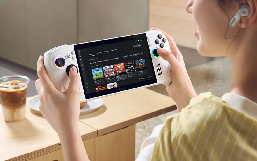 Persoon speelt op witte ROG Xbox Ally handheld console, zittend aan houten tafel met koffie, draagt gele trui en oordopjes.