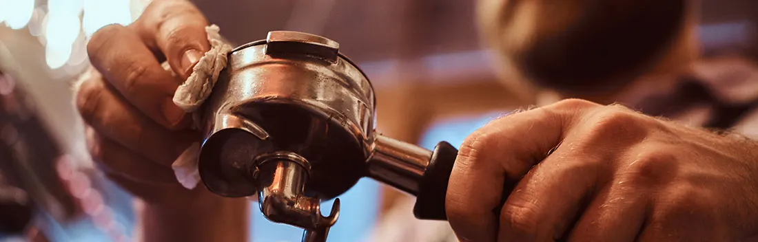 Een zilverkleurige koffiefilterhouder wordt schoongemaakt met een doek. De achtergrond is wazig.