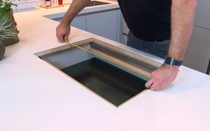 Plan de travail blanc avec découpe rectangulaire mesurée par un mètre ruban jaune, fond de cuisine avec armoires grises.
