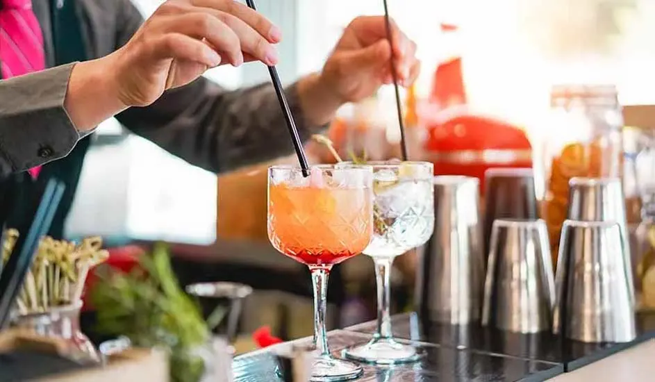 Deux cocktails dans des verres à pied, orange et transparent, avec pailles noires. Shakers en métal en arrière-plan.