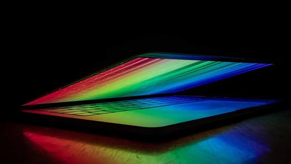 A half-closed Apple MacBook radiates bright rainbow colours onto keyboard and table; Apple design recognisable.