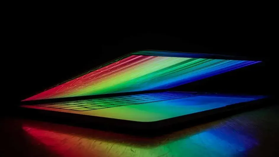 A half-closed Apple MacBook radiates bright rainbow colours onto keyboard and table; Apple design recognisable.