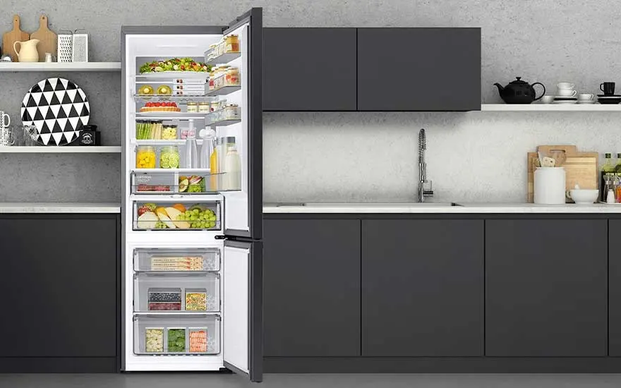 Un réfrigérateur ouvert, rempli de divers aliments, est placé dans une cuisine moderne aux armoires sombres et plans de travail clairs.