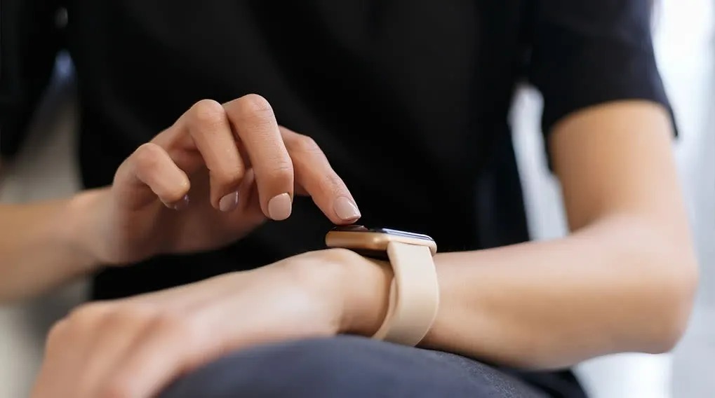 Een hand tikt op een smartwatch met een beige band tegen een achtergrond van een zwart shirt.