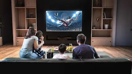 Een woonkamer met een zwart tv-toestel aan de muur, houten kasten en een zwarte sofa. Een tafel met fruit en sap staat voor de sofa.