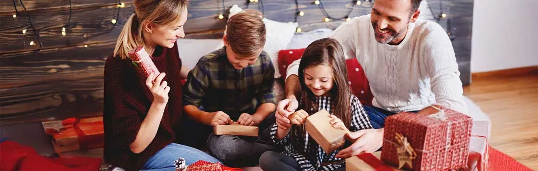 Une famille est confortablement réunie sur le canapé, ouvrant joyeusement des cadeaux de Noël, entourée de guirlandes lumineuses.