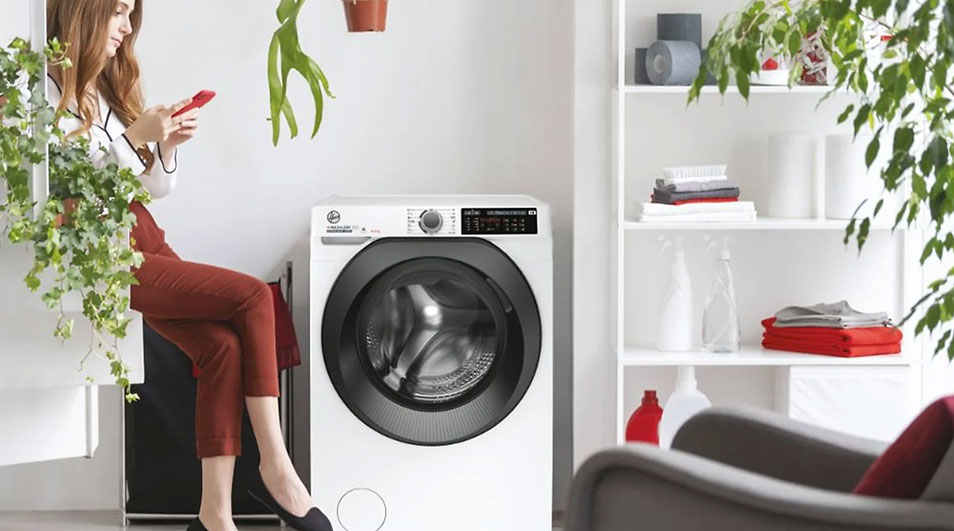 A white washing machine with a black door is in a room with a white shelf holding folded clothes and bottles. A plant hangs above.