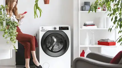 A white washing machine with a black door is in a room with a white shelf holding folded clothes and bottles. A plant hangs above.