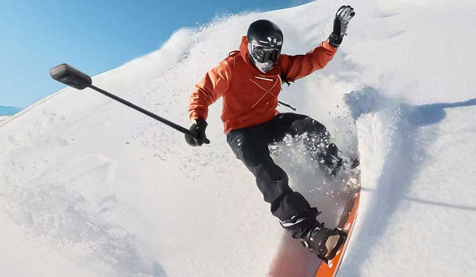 Un snowboardeur en veste orange se filme avec une GoPro sur perche, glissant rapidement dans la neige fraîche sur une montagne.