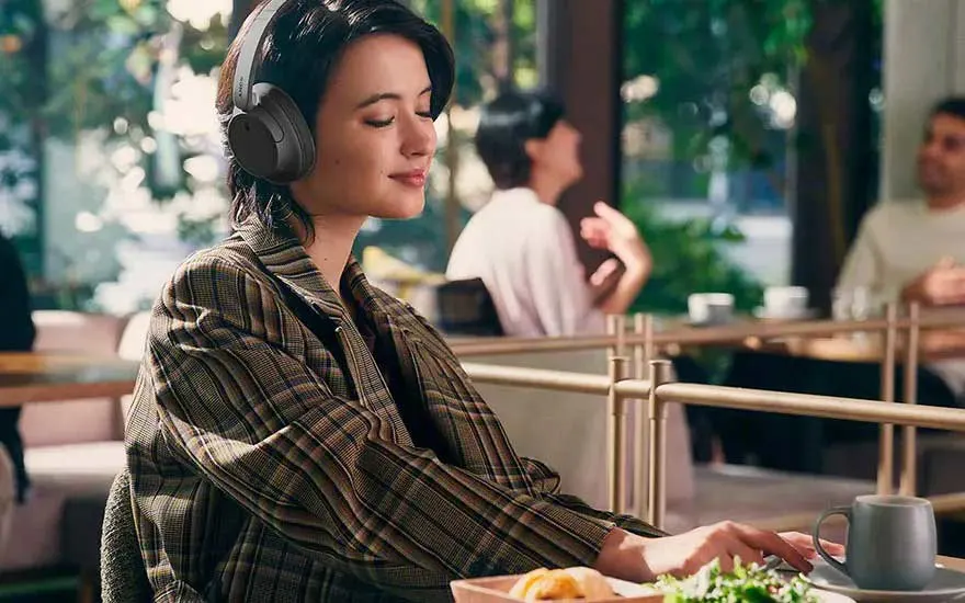 Une personne avec un casque audio porte une veste à carreaux. Devant, il y a de la nourriture et une tasse. En arrière-plan, d'autres personnes sont assises.