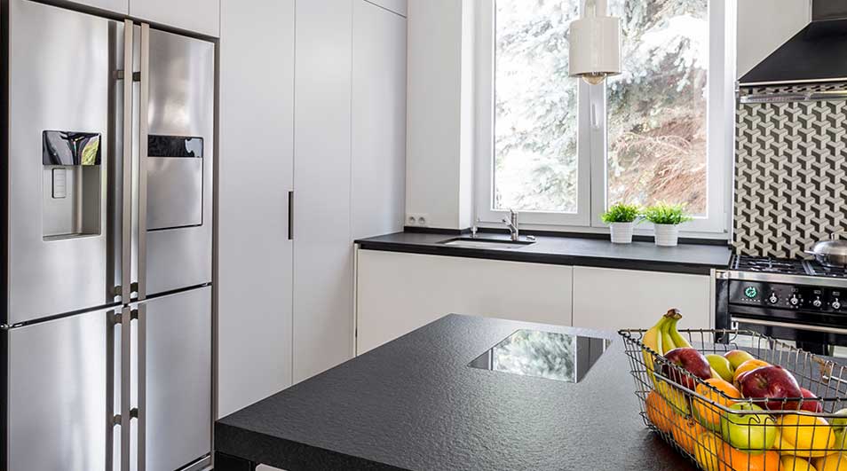 Cuisine moderne avec frigo en inox, îlot noir, fenêtre et fruits dans un panier en métal. Crédence à motifs géométriques.