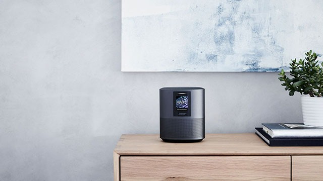 Haut-parleur gris sur une commode en bois clair, près d'un tableau et d'une plante verte en pot blanc. Mur gris clair en arrière-plan.