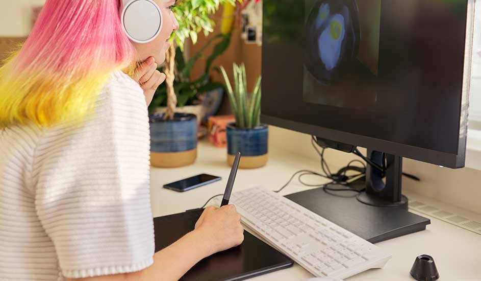 Une tablette graphique noire, un stylet, un clavier blanc et un écran d'ordinateur noir sur un bureau blanc. Des plantes en arrière-plan.
