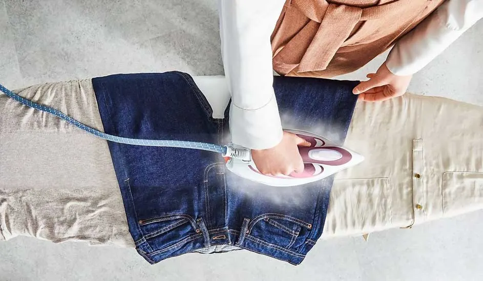 Een blauwe jeans wordt gestreken op een strijkplank met een beige shirt. Er komt stoom uit het strijkijzer.