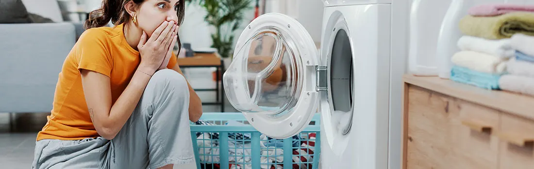 Une machine à laver blanche ouverte, un panier à linge bleu et une pile de serviettes sur une commode en bois.