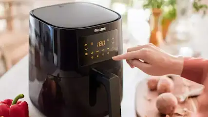 Un friteuse à air noire Philips affiche 180 degrés. Une main sélectionne des réglages. Un poivron rouge et un plat avec des aliments sont visibles.