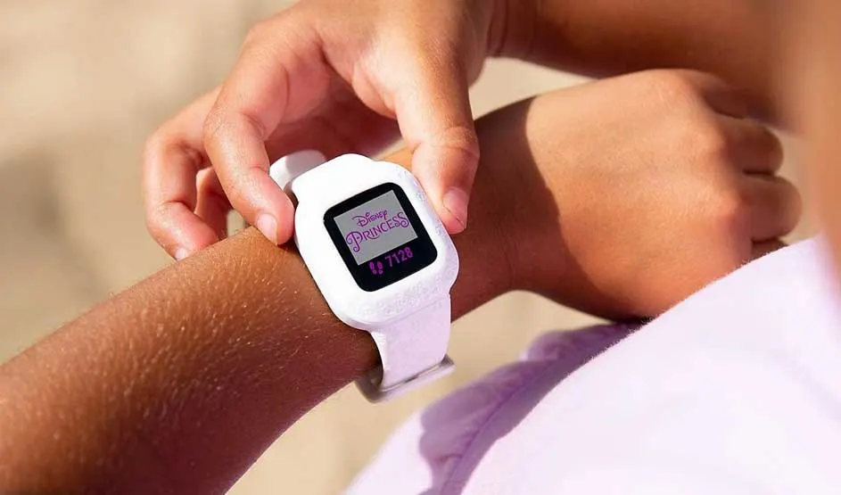 Een kind draagt een witte smartwatch met Disney Princess-scherm; met de andere hand raakt het kind het horloge aan in warm licht.