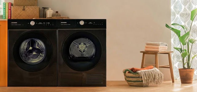 Deux machines à laver noires, un tabouret en bois, un panier et une plante dans une pièce lumineuse.