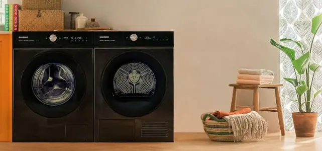 Zicht op een wasmachine en droogkast in het zwart, naast een houten kruk met handdoeken en een plant in een pot.