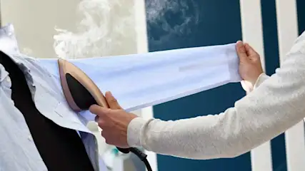 Een blauw overhemd wordt gestoomd met een stoomstrijkijzer, er komt stoom uit. Op de achtergrond een blauw-witte muur.
