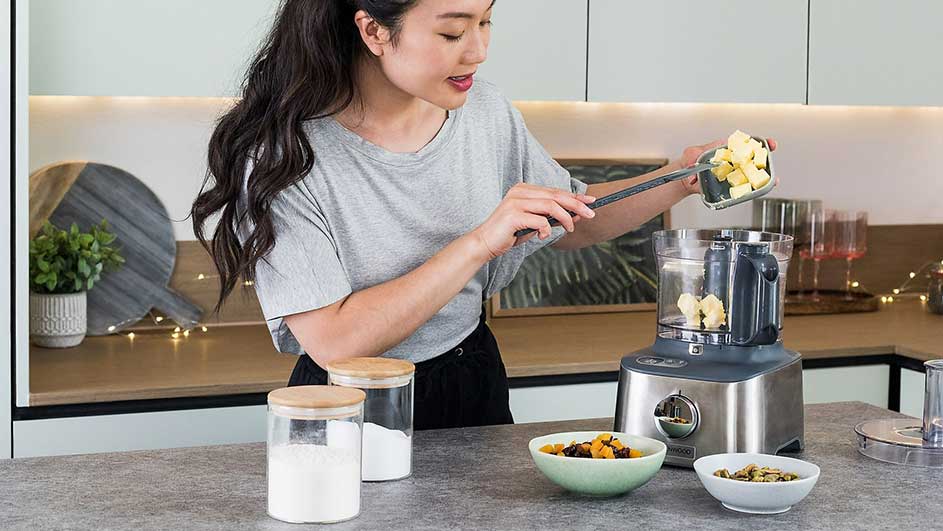 Keuken scène: Een grijze keukenmachine met een doorzichtige kom, gevuld met stukjes fruit. Schalen met ingrediënten en glazen potten staan klaar op het aanrecht.