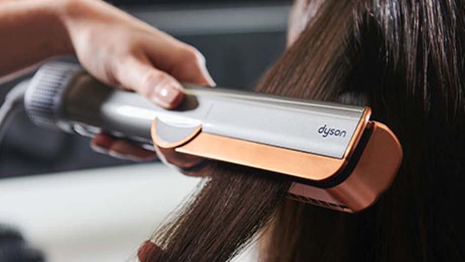 Une main tient un fer à lisser Dyson gris et bronze sur des cheveux bruns.