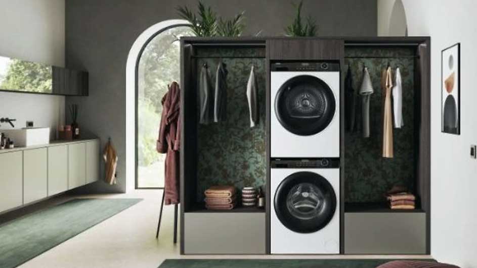 Witte wasmachine en droger gestapeld in kast met hangende kleding. Een wastafel en een raam zijn zichtbaar aan de linkerkant.