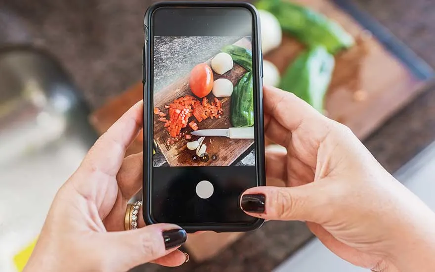 Gros plan sur des mains prenant en photo une planche à découper avec légumes et couteau, la mise au point est sur l’écran du smartphone.