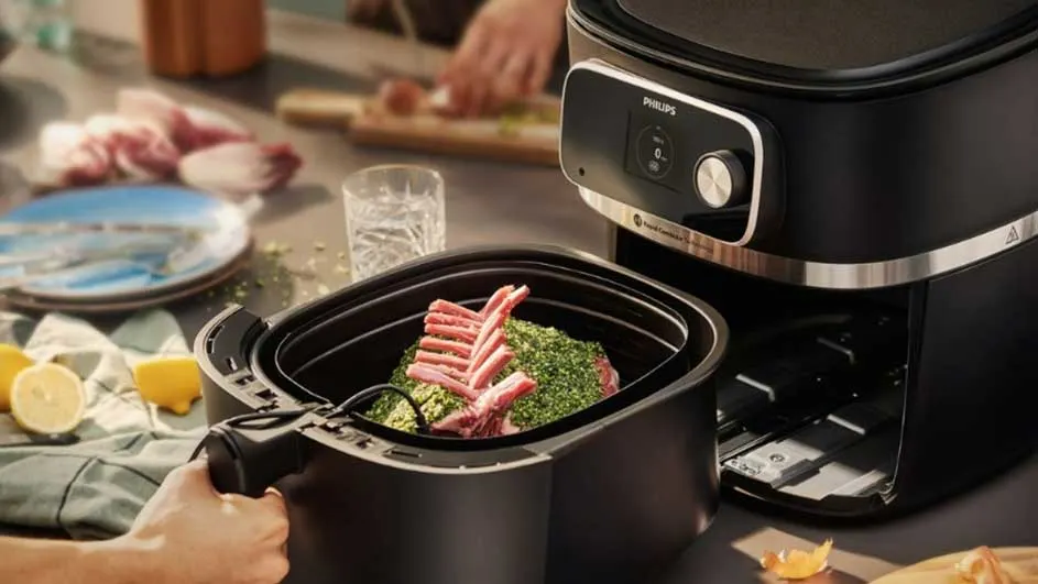 Vue rapprochée d'une friteuse à air Philips noire avec un carré d'agneau dans le panier. Citrons et assiettes en arrière-plan.