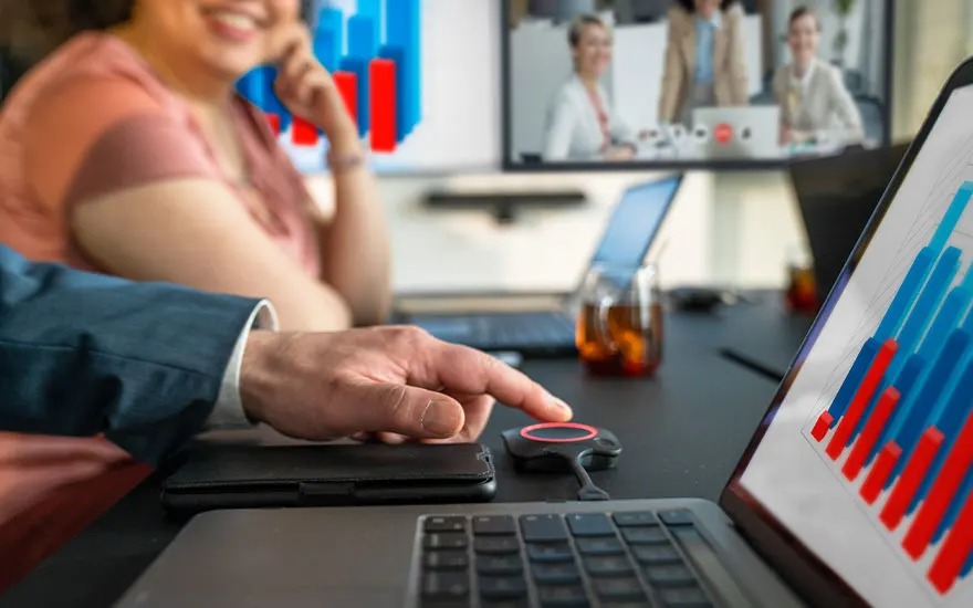 Een hand drukt op een ClickShare-knop naast een laptop met staafdiagram, collega’s glimlachen en hebben een online meeting op scherm.