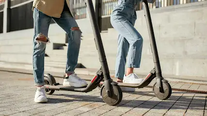 Twee personen op elektrische steps op een betegelde ondergrond. De steps zijn donkergrijs en de personen dragen jeans en witte schoenen.