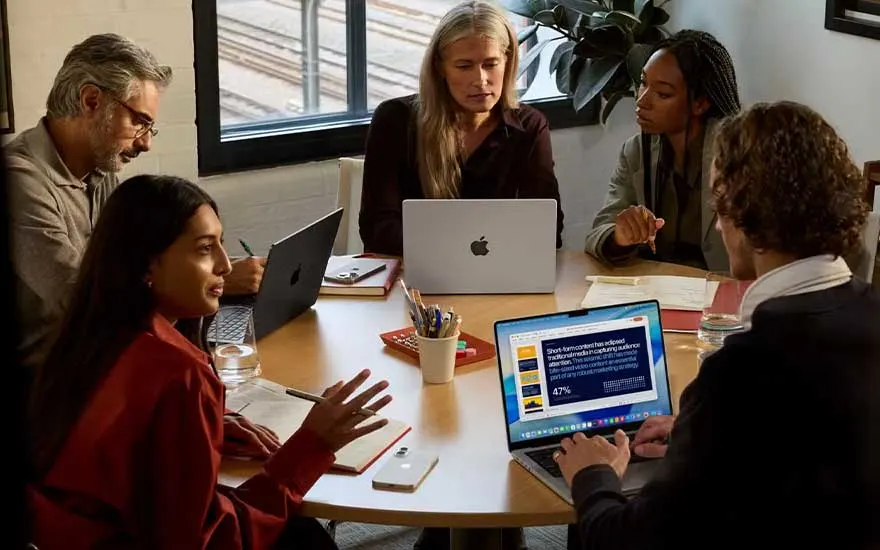 Cinq collègues échangent autour d’une table avec ordinateurs et notes, pendant qu’une présentation s’affiche à l’écran.
