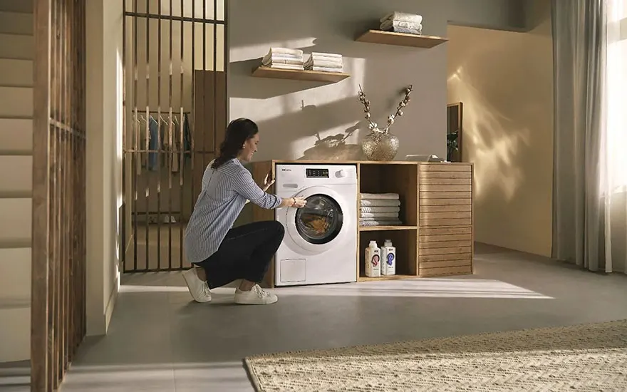 Un lave-linge blanc à côté d'étagères en bois dans une pièce. Des serviettes blanches sont pliées sur les étagères.