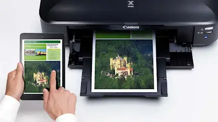 Une imprimante Canon noire imprime une photo de château. Une tablette affiche la même image. Mains visibles.