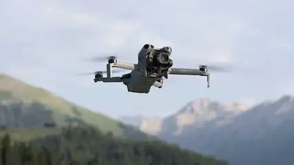 This is a gray DJI Mini 3 Pro drone hovering in the air above mountains, with a camera at the front.