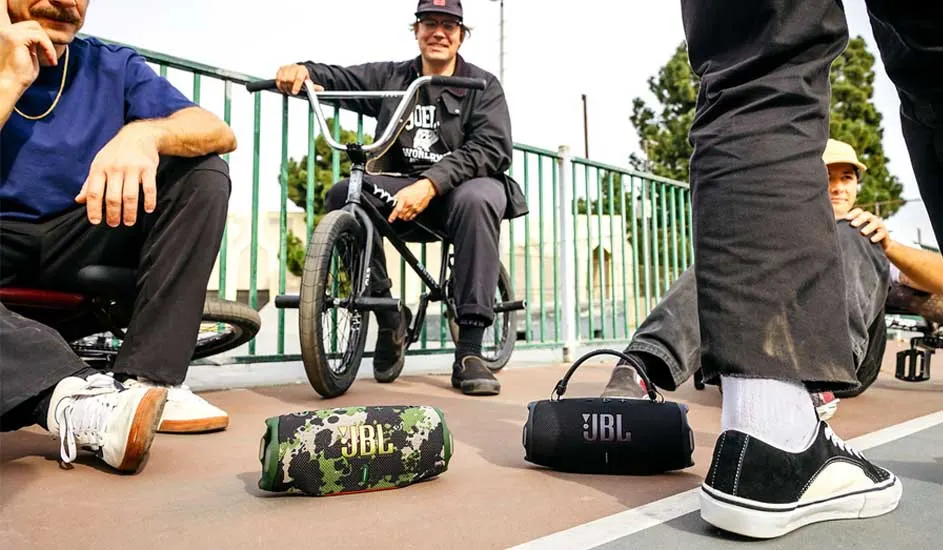 Des haut-parleurs JBL, un vélo BMX et des personnes sont visibles sur une surface grise avec une clôture verte en arrière-plan.