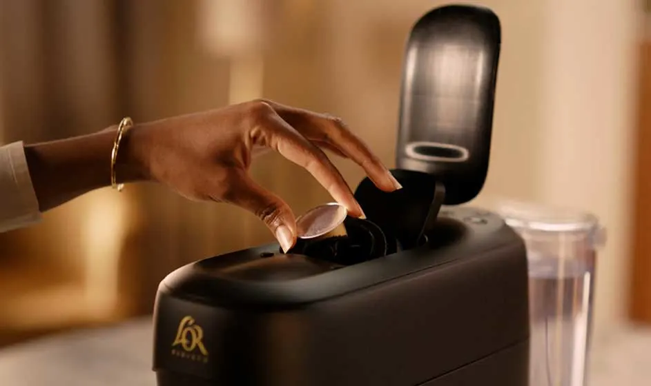 Een hand plaatst een capsule in een zwarte L'OR-koffiemachine, met open klep en waterreservoir in zacht, warm keukenlicht.