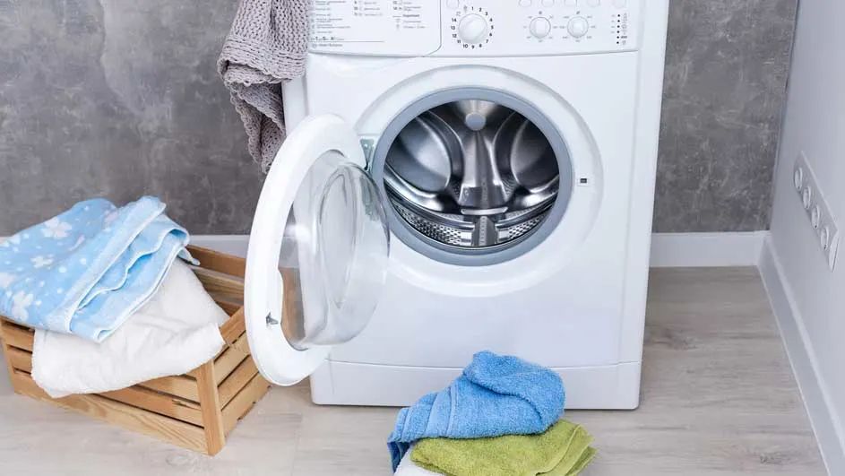 Witte wasmachine met open deur, houten bak met handdoeken en stapel handdoeken op de vloer. Grijze muur op achtergrond.