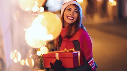 Een vrouw met een kerstmuts loopt vrolijk langs feestelijke lichtjes buiten en houdt een rood, goud gestrikt cadeautje vast.