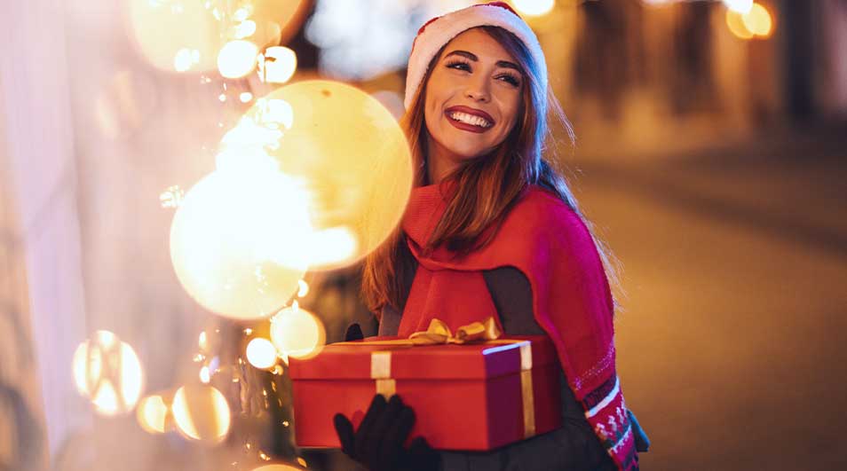 Une femme portant un bonnet de Noël marche près de lumières festives, tenant une boîte cadeau rouge nouée d’un ruban doré.