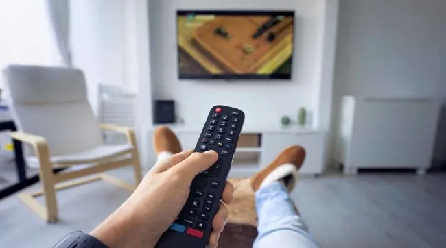 Een hand houdt een zwarte afstandsbediening vast voor een tv in een lichte woonkamer met stoelen.