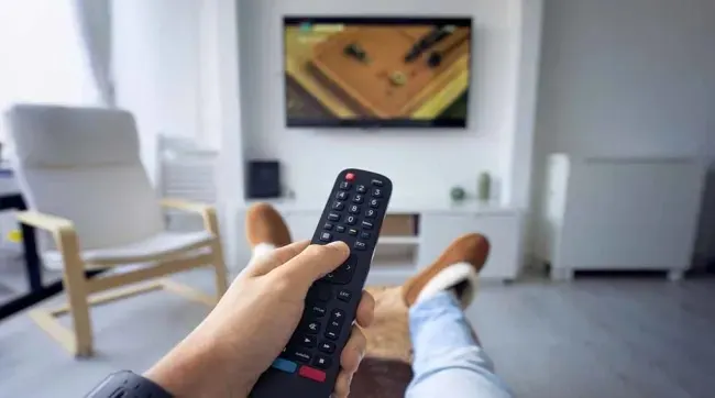 Een hand houdt een zwarte afstandsbediening vast voor een tv in een lichte woonkamer met stoelen.