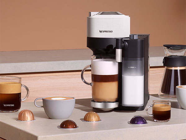 Een Nespresso-apparaat met koffiekopjes, capsules en een glazen koffiekan op een witte tafel tegen een perzikachtige achtergrond.