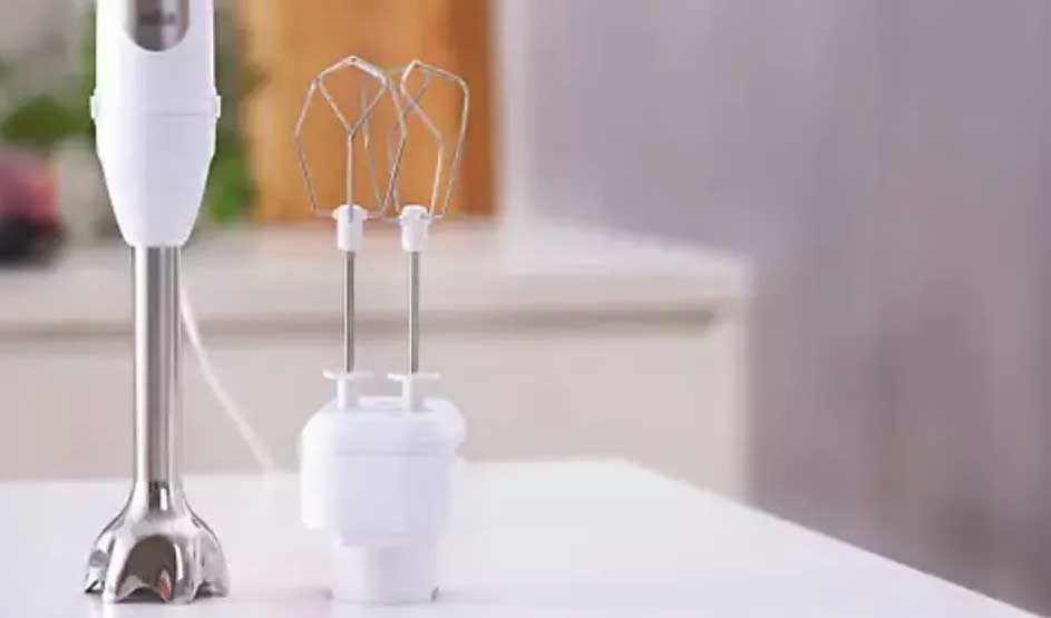 Een witte staafmixer en kloppers staan op een wit aanrechtblad. Achtergrond is een keuken.